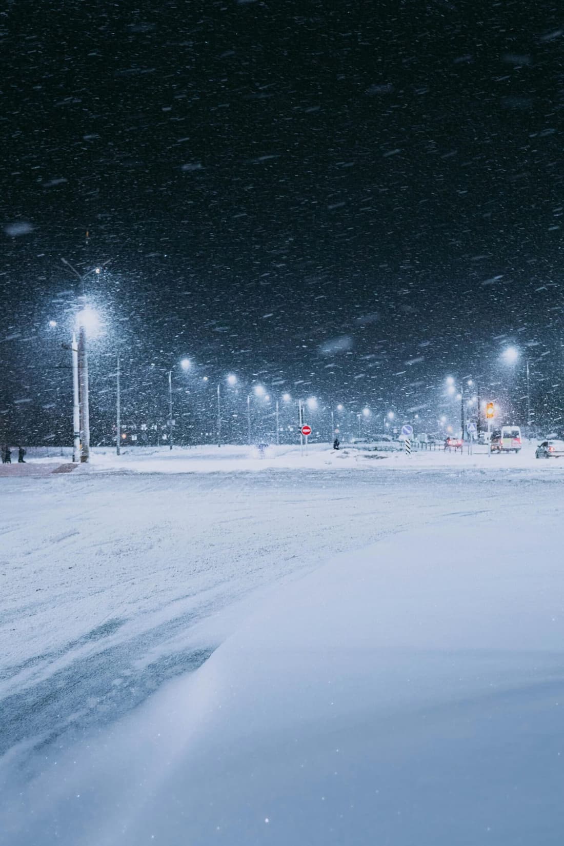 A snowy city street at night with heavy snowfall, illuminated by streetlights and traffic signals.