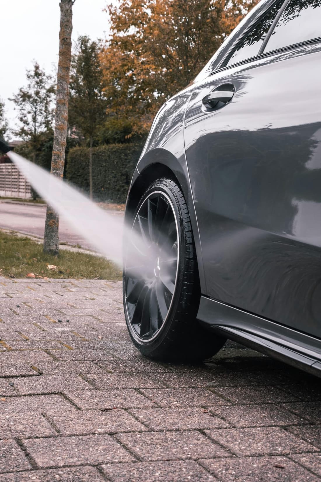 Close-up of a sleek car being pressure washed, focusing on the clean black alloy wheel and reflecting the autumn surroundings.