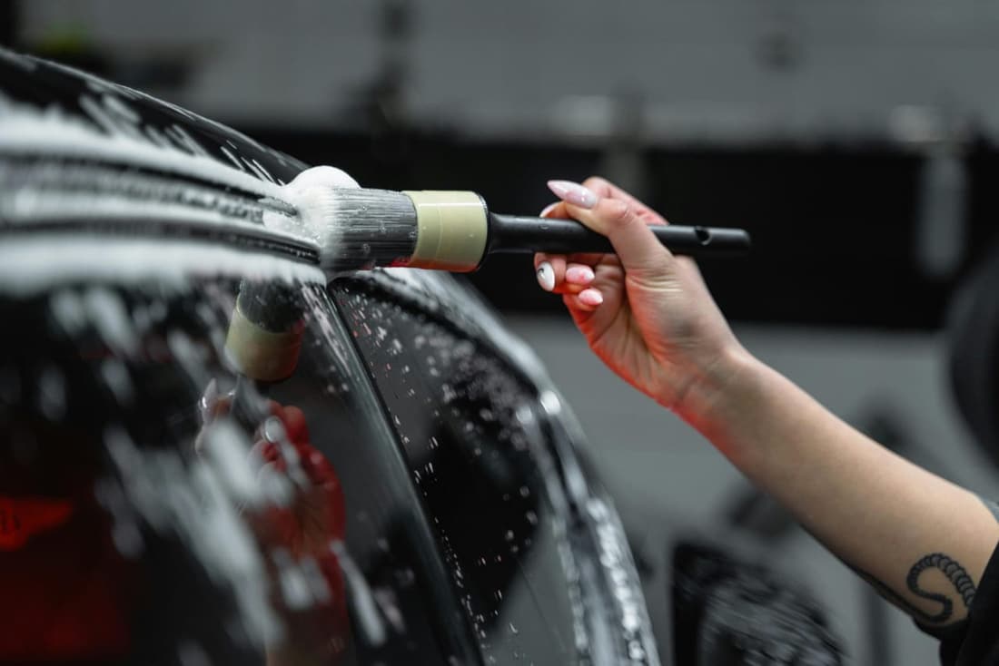 Close-up of a professional detailer using a brush to clean a soapy car exterior, ensuring precision and shine during the detailing process.