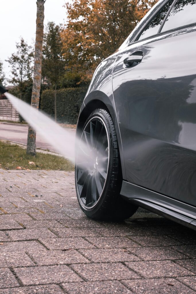 Close-up of a sleek car being pressure washed, focusing on the clean black alloy wheel and reflecting the autumn surroundings.