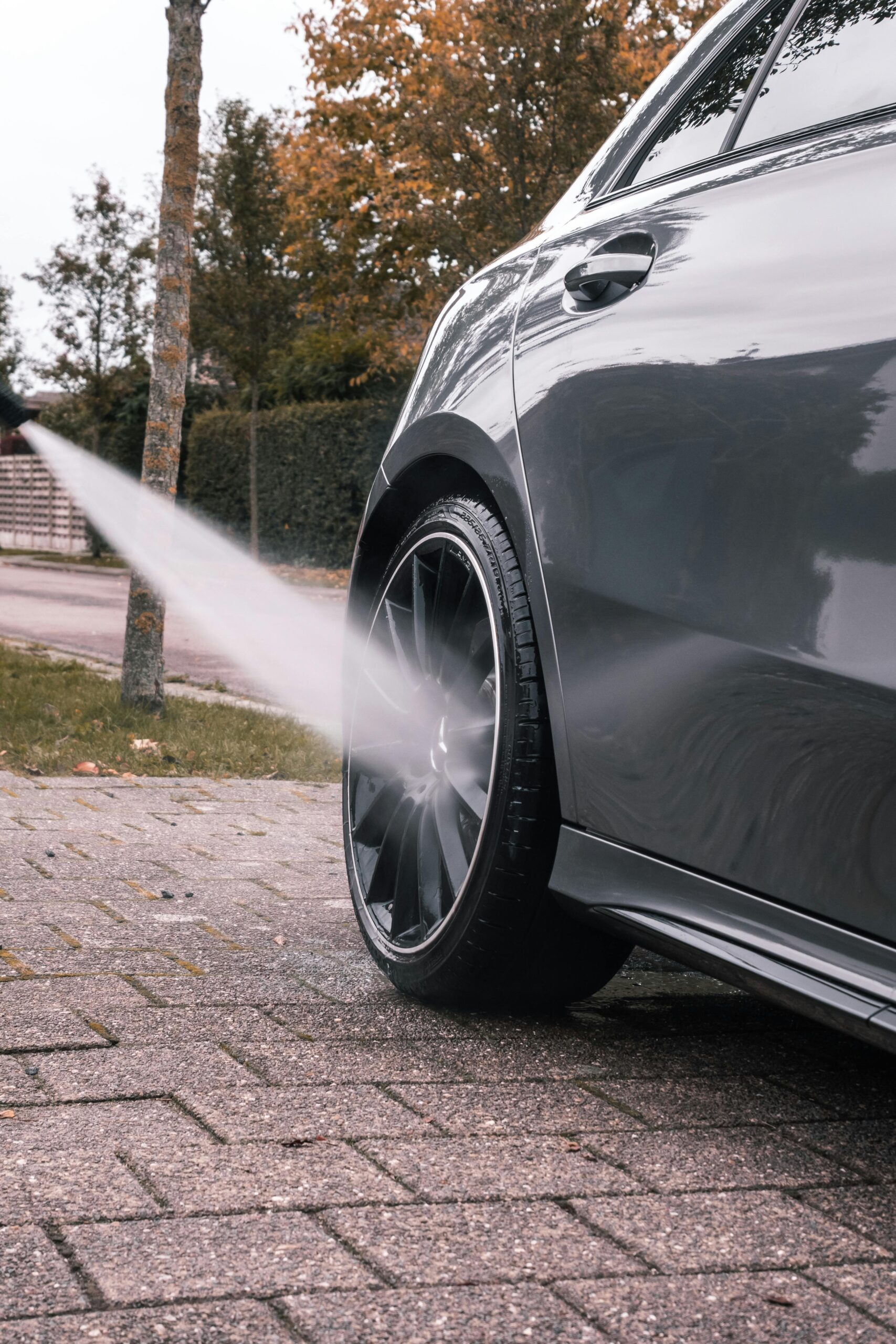 pexels-jarne-aerts-3175149-5693660 Close-up of a sleek car being pressure washed, focusing on the clean black alloy wheel and reflecting the autumn surroundings.
